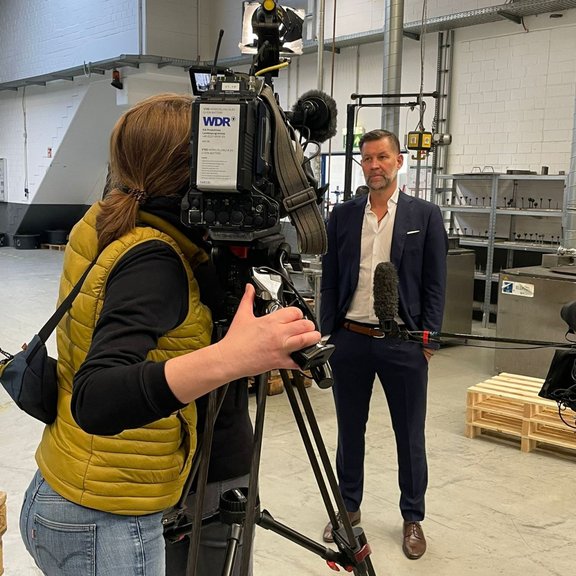 Auf dem Bild ist eine Kamerfrau zu sehen, die Geschäftsführer Marco Dörr für einen Fernsehbeitrag aufnimmt. Sie stehen in der Produktionshalle von Stannol.
