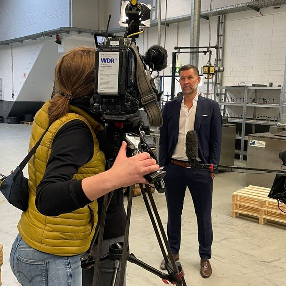 The picture shows a camerawoman filming Managing Director Marco Dörr for a television programme. They are standing in the Stannol production hall.