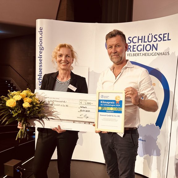 The picture shows Managing Director Marco Dörr and Management Assistant Susanne Schlüter. They are holding a large cheque, a bouquet of flowers and a certificate in their hands and smiling friendly into the camera. A display of the key region can be seen in the background.